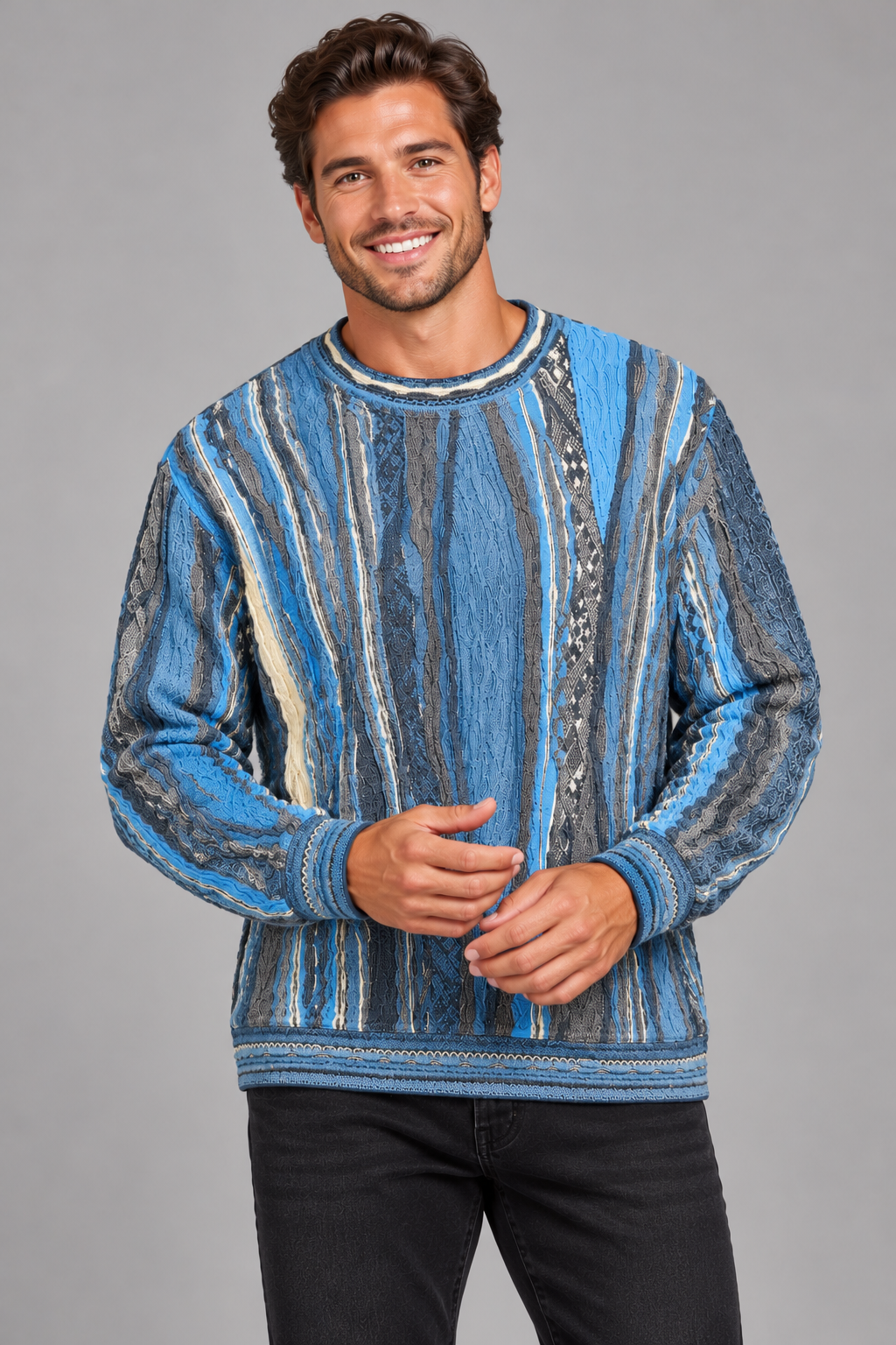 A man wearing a blue and gray vertically striped knit sweater with textured patterns, paired with black jeans, standing against a solid gray studio background.
