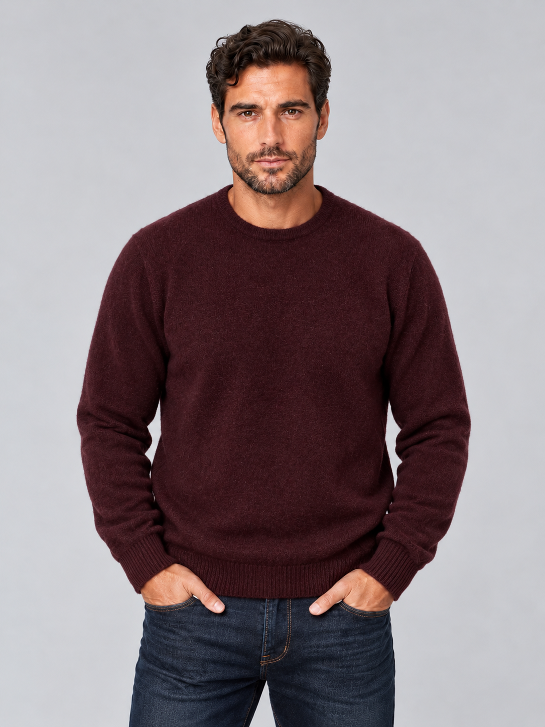 A man wearing a dark burgundy crewneck sweater and dark blue jeans, standing against a solid gray studio background.