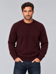A man wearing a dark burgundy crewneck sweater and dark blue jeans, standing against a solid gray studio background.