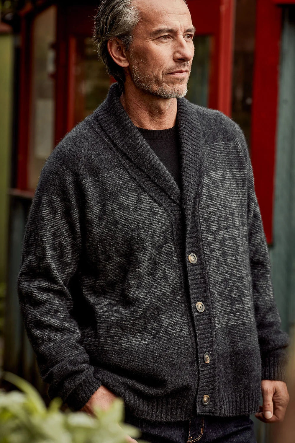 A man wearing a dark charcoal gray patterned knit cardigan over a black shirt, standing outdoors in front of a red building.