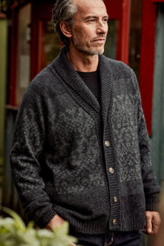 A man wearing a dark charcoal gray patterned knit cardigan over a black shirt, standing outdoors in front of a red building.