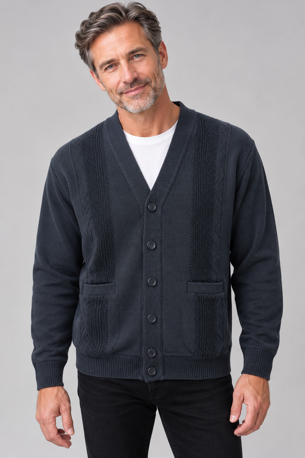 A man wearing a dark navy blue button-up cardigan sweater with textured knit panels, paired with a white t-shirt and black pants, against a solid gray studio background.