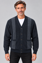 A man wearing a dark navy blue cardigan sweater with textured gray knit shoulder and front panel details, layered over a white t-shirt and black pants, against a solid gray studio background.