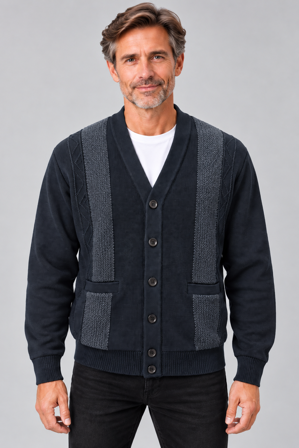 A man wearing a dark navy blue cardigan sweater with textured gray knit shoulder and front panel details, layered over a white t-shirt and black pants, against a solid gray studio background.