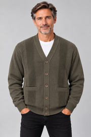 A man wearing an olive green knit cardigan with textured panels and button-front closure, layered over a white t-shirt and black pants, against a solid gray studio background.