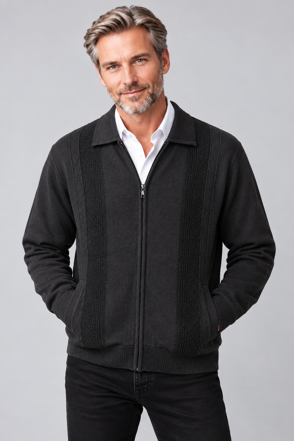 A man wearing a black zip-up jacket with textured knit panels on the chest, worn over a white collared shirt and black pants, against a solid gray studio background.