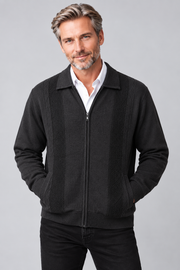 A man wearing a black zip-up jacket with textured knit panels on the chest, worn over a white collared shirt and black pants, against a solid gray studio background.
