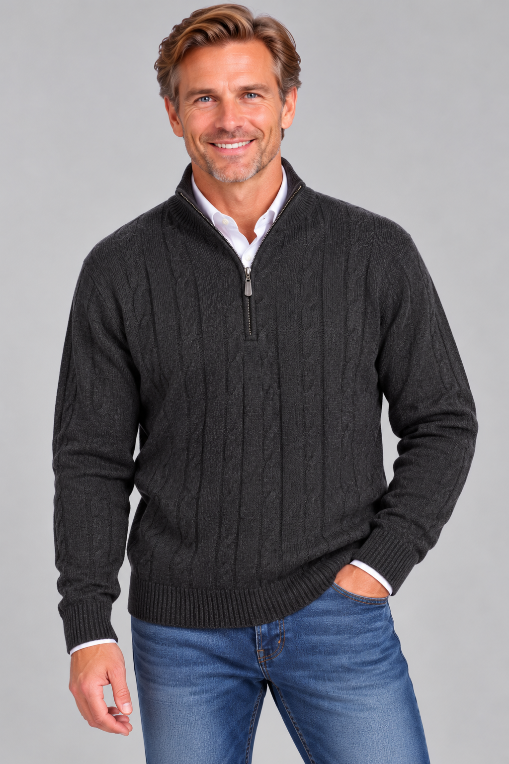 A smiling man wearing a dark charcoal gray cable-knit quarter-zip sweater over a white collared shirt and blue jeans, standing against a solid gray studio background.