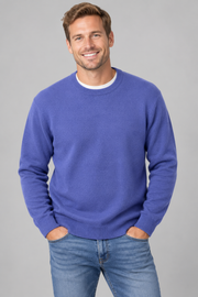 A smiling man wearing a plum blue crewneck sweater over a white undershirt and blue jeans, standing against a solid grey studio background.