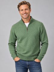 A smiling man wearing a green quarter-zip knit sweater over a white t-shirt and blue jeans, standing against a solid gray studio background.