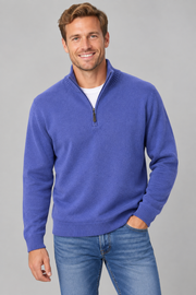A smiling man wearing a plum blue quarter-zip knit sweater over a white t-shirt and blue jeans, standing against a solid grey studio background.
