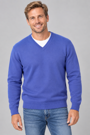 A smiling man wearing a royal blue v-neck knit sweater over a white undershirt and blue jeans, photographed against a plain light grey studio background.