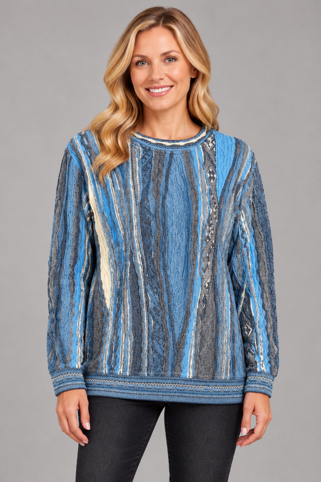 A woman modeling a long-sleeve crewneck sweater with a vertical striped pattern in shades of blue, gray, and beige, paired with black pants against a solid gray studio background.