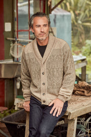 A man wearing a textured beige knit cardigan over a black shirt and dark jeans, sitting on a rustic wooden bench in an outdoor workshop setting.