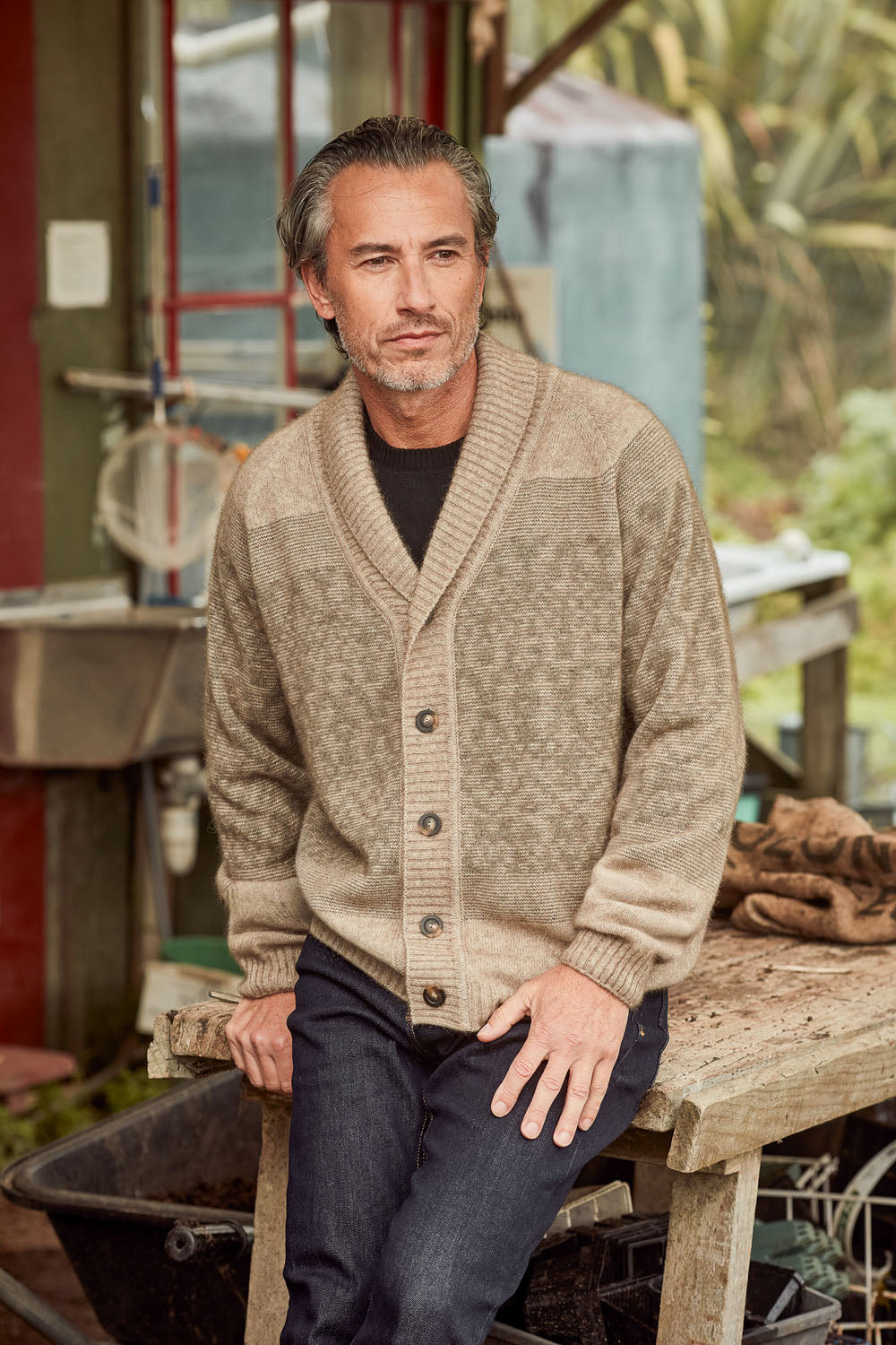 A man wearing a textured beige knit cardigan over a black shirt and dark jeans, sitting on a rustic wooden bench in an outdoor workshop setting.