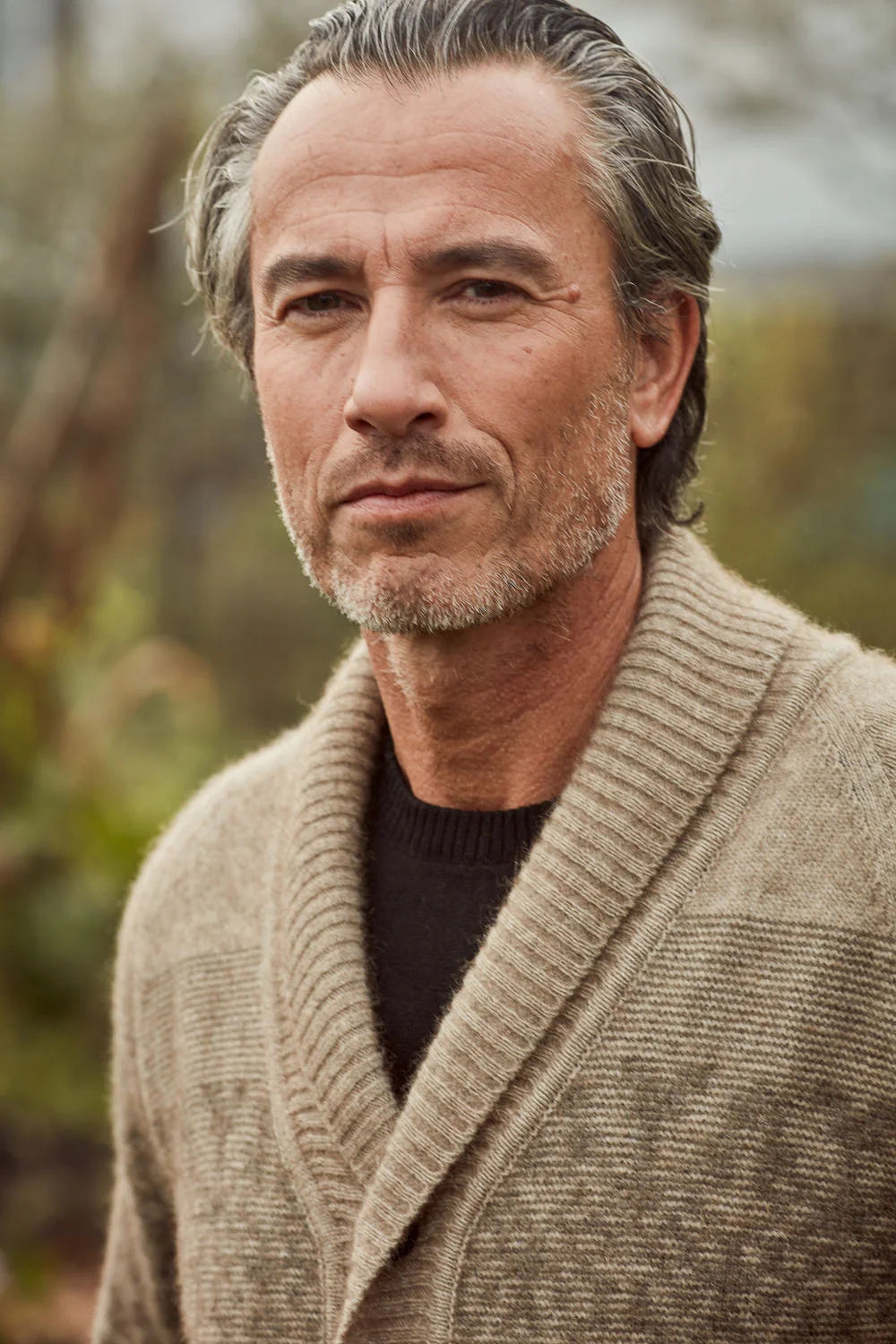 A close-up portrait of a man wearing a textured beige shawl-collar knit cardigan over a black crewneck sweater, set against a soft, blurred outdoor background.