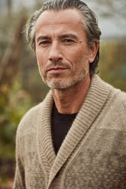 A close-up portrait of a man wearing a textured beige shawl-collar knit cardigan over a black crewneck sweater, set against a soft, blurred outdoor background.