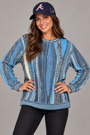 A woman wearing a blue and gray vertically striped knit sweater with a crew neck, paired with black jeans and a navy baseball cap with a white 'A' logo, posed against a solid gray studio background.