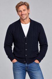 A smiling man wearing a dark navy blue cable-knit cardigan over a white t-shirt and blue jeans, standing against a solid gray studio background.