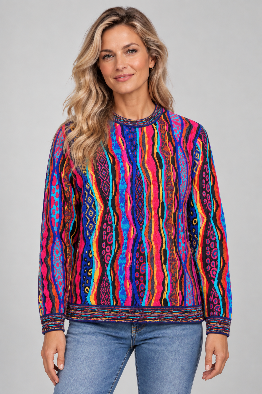 A woman wearing a vibrant, multicolored long-sleeve crewneck sweater with vertical wavy stripes in pink, blue, orange, yellow, and purple, paired with blue jeans, against a solid gray studio background.
