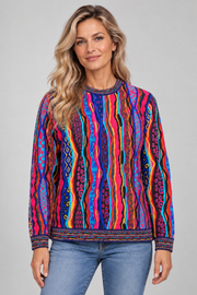 A woman wearing a vibrant, multicolored long-sleeve crewneck sweater with vertical wavy stripes in pink, blue, orange, yellow, and purple, paired with blue jeans, against a solid gray studio background.