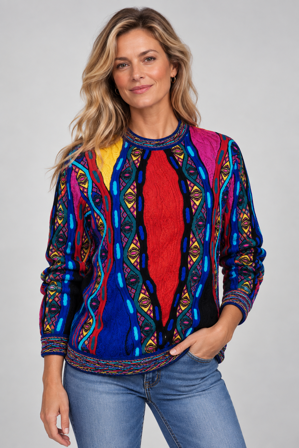 A woman modeling a vibrant, multicolored long-sleeve knit sweater with intricate geometric and tribal-inspired patterns in blue, red, yellow, pink, and black, paired with blue jeans against a solid light gray background.