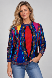 A woman modeling a vibrant, multicolored long-sleeve knit sweater with intricate geometric and tribal-inspired patterns in blue, red, yellow, pink, and black, paired with blue jeans against a solid light gray background.
