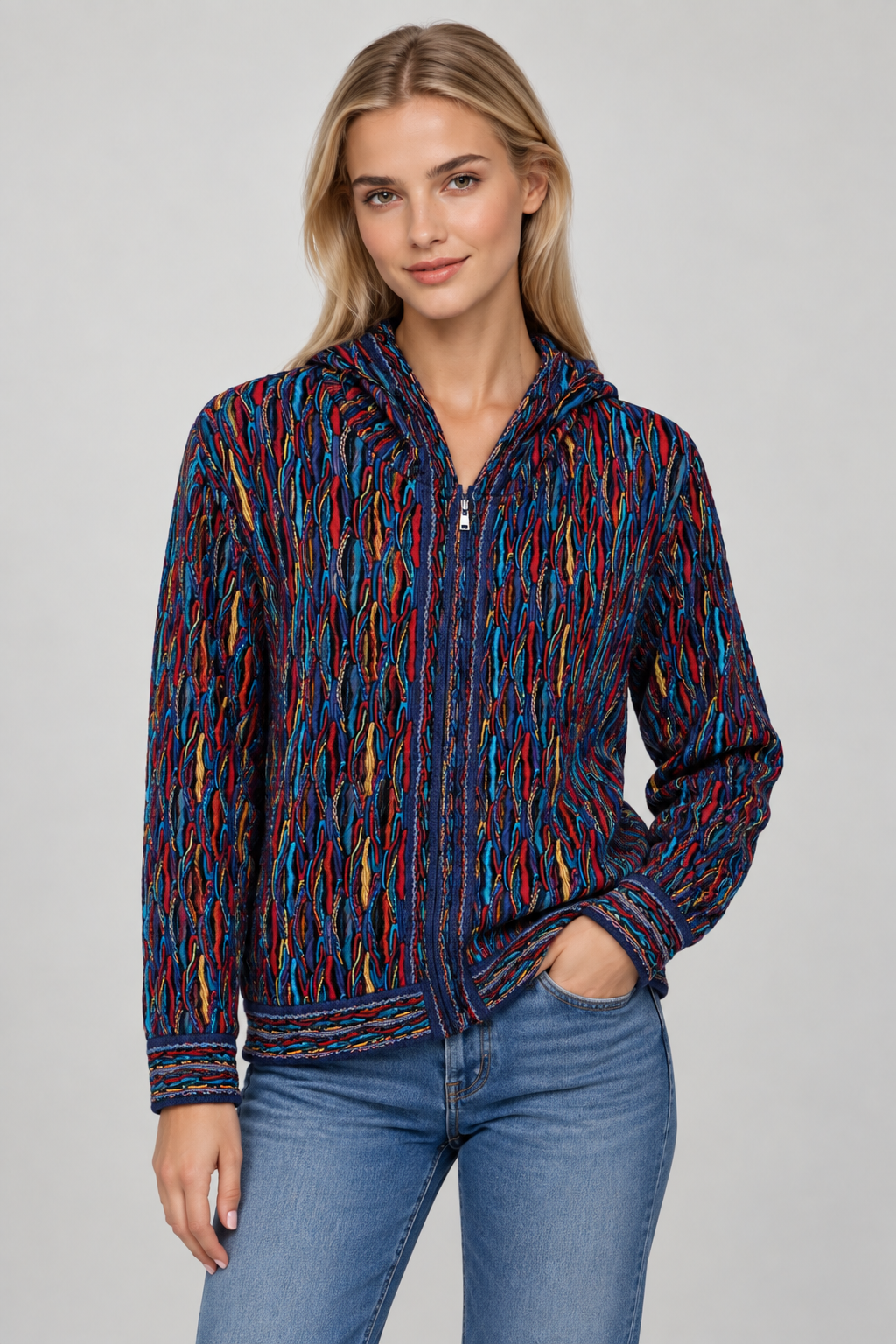 A woman modeling a zip-up hooded jacket with a multicolor abstract knit pattern in blue, red, orange, and yellow, paired with blue jeans against a light gray studio background.