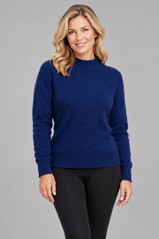 A woman modeling a navy blue ribbed knit mock neck sweater with black leggings, standing against a solid gray studio background.