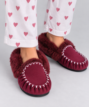 A lady wearing burgundy sheepskin-fur slippers with white contrast stitching, paired with white pyjama pants printed with pink hearts, on a light grey studio background.
