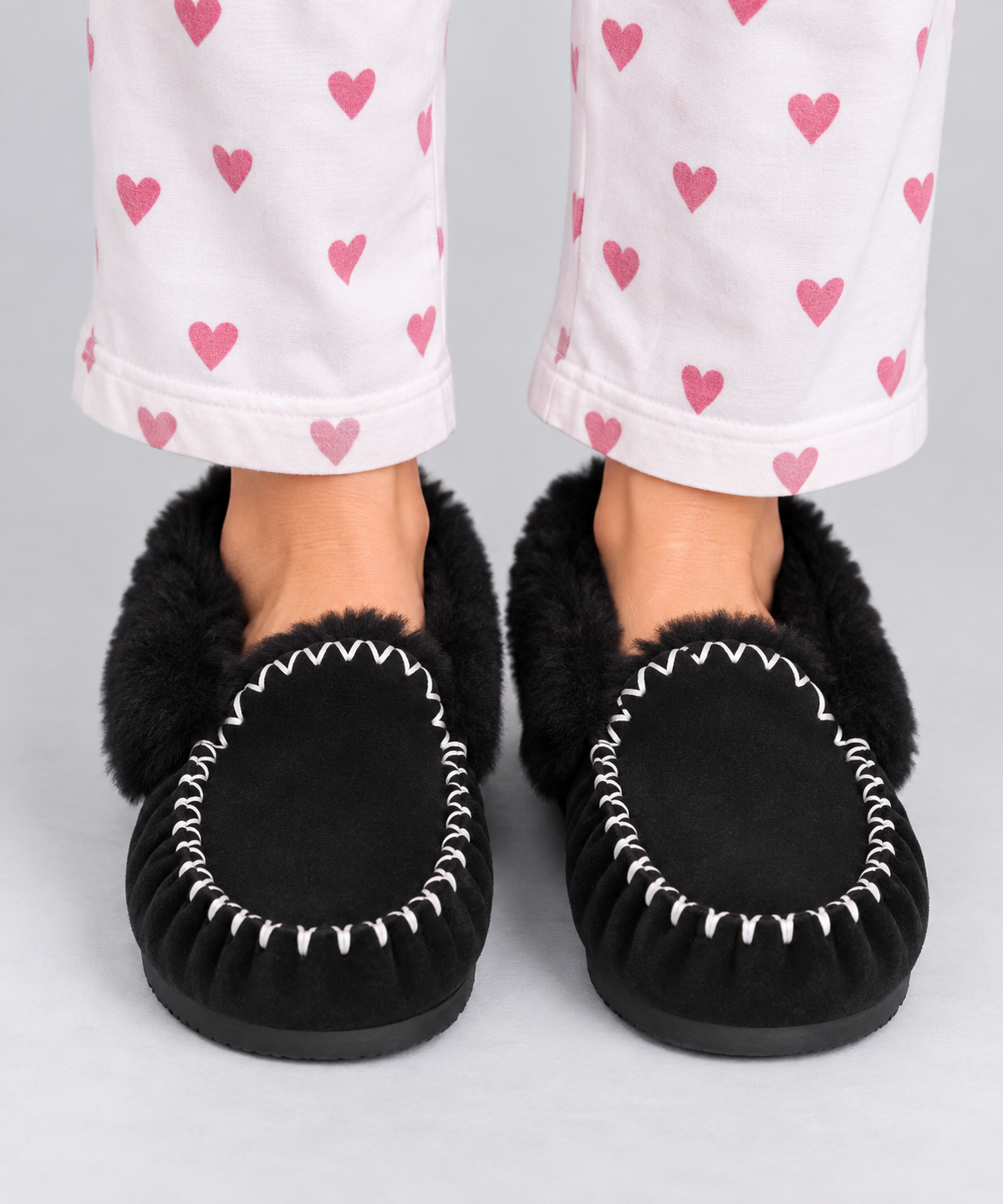Black suede moccasin slippers with black 100% sheepskin fur and white decorative stitching, shown on feet with white pyjama pants printed with pink hearts.