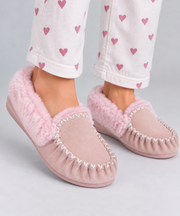 A person wearing pink suede-like slip-on slippers with fluffy pink lining and white decorative stitching, paired with white pants printed with pink hearts, on a light gray studio background.