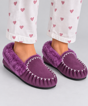 Purple suede moccasin slipper with a 100% genuine sheepskin fur internal and trim, shown on a person's feet wearing white pants with a pink heart pattern, against a light gray background.