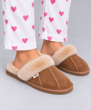 A person wearing tan suede slip-on slippers with fluffy cream sheepskin fur, paired with white pants printed with pink hearts, against a light grey studio background.
