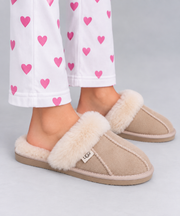 A person wearing beige UGG slip-on slippers with fluffy cream sherpa trim and white pants with pink heart prints, shown against a light gray studio background.