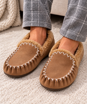 A person wearing tan suede moccasin-style slippers with fluffy brown lining and white decorative stitching, shown with gray plaid pants on a light carpet.