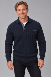 A smiling man wearing a dark navy ribbed knit polo sweater over a light blue collared shirt and black pants, standing against a solid gray studio background.