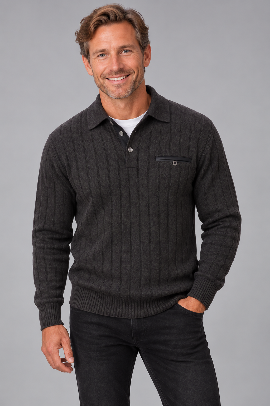 A smiling man wearing a dark charcoal gray ribbed knit polo sweater over a white undershirt and black pants, standing against a solid gray studio background.