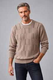 A man wearing a beige textured knit crewneck sweater with a basketweave pattern on the chest, paired with dark blue jeans, against a solid gray studio background.