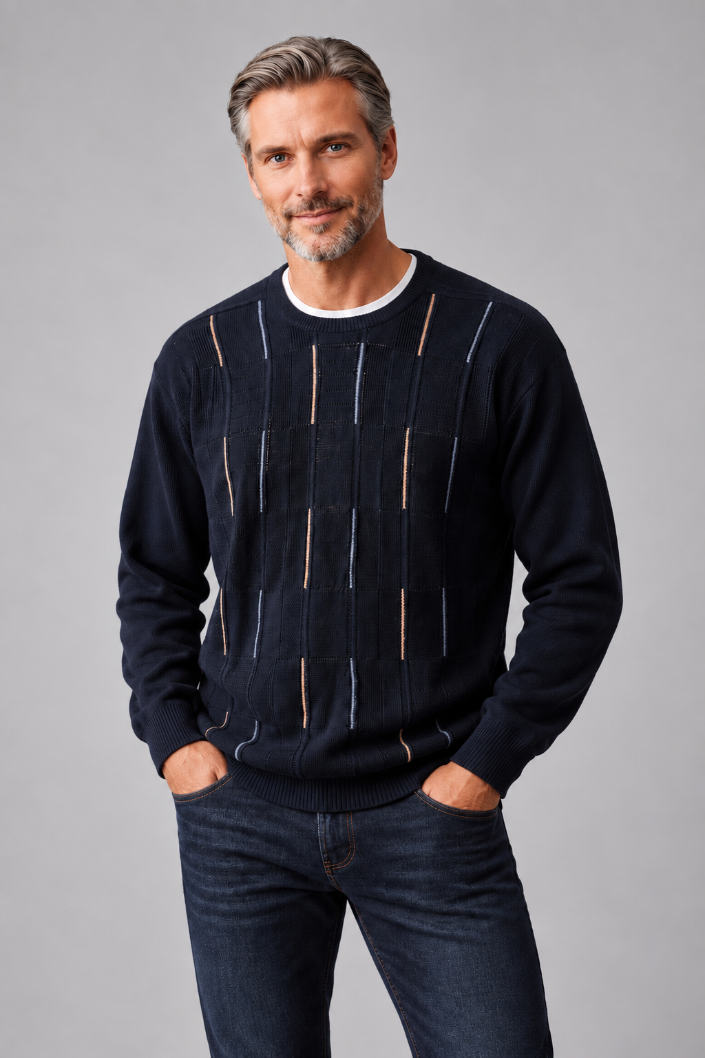 A man wearing a dark navy blue crewneck sweater with vertical striped knit patterns in beige and white, paired with dark blue jeans, against a solid gray studio background.