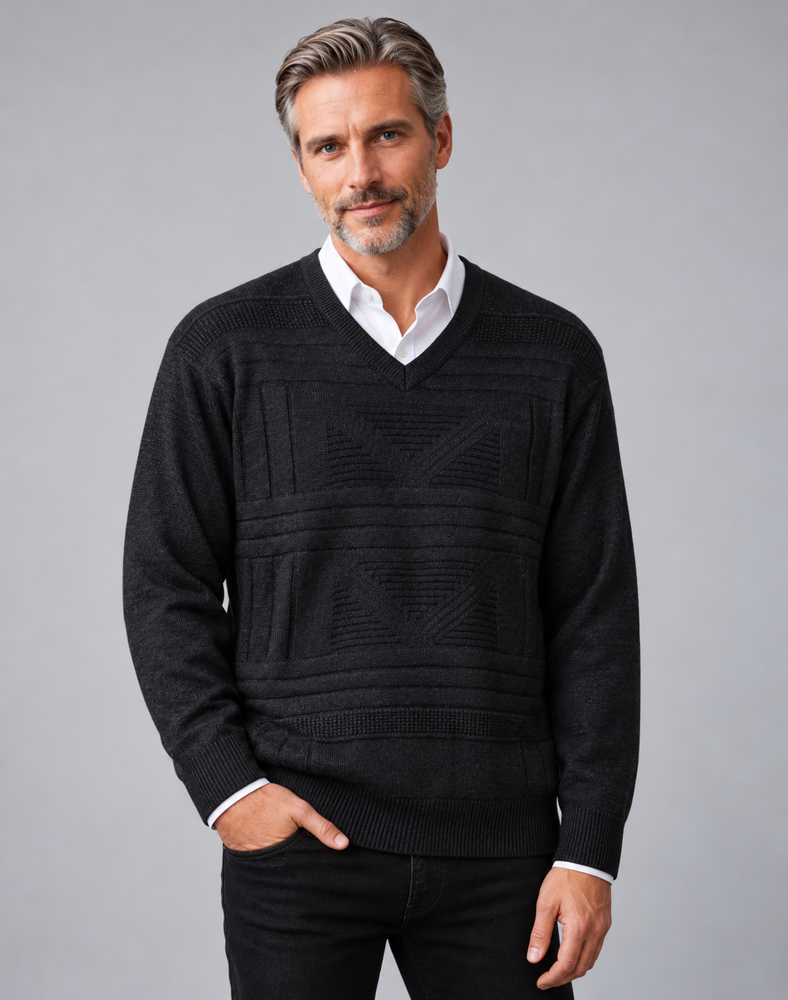 A man wearing a black textured knit v-neck sweater over a white collared shirt, paired with black trousers, photographed against a solid gray studio background.