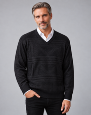 A man wearing a black textured knit v-neck sweater over a white collared shirt, paired with black trousers, photographed against a solid gray studio background.