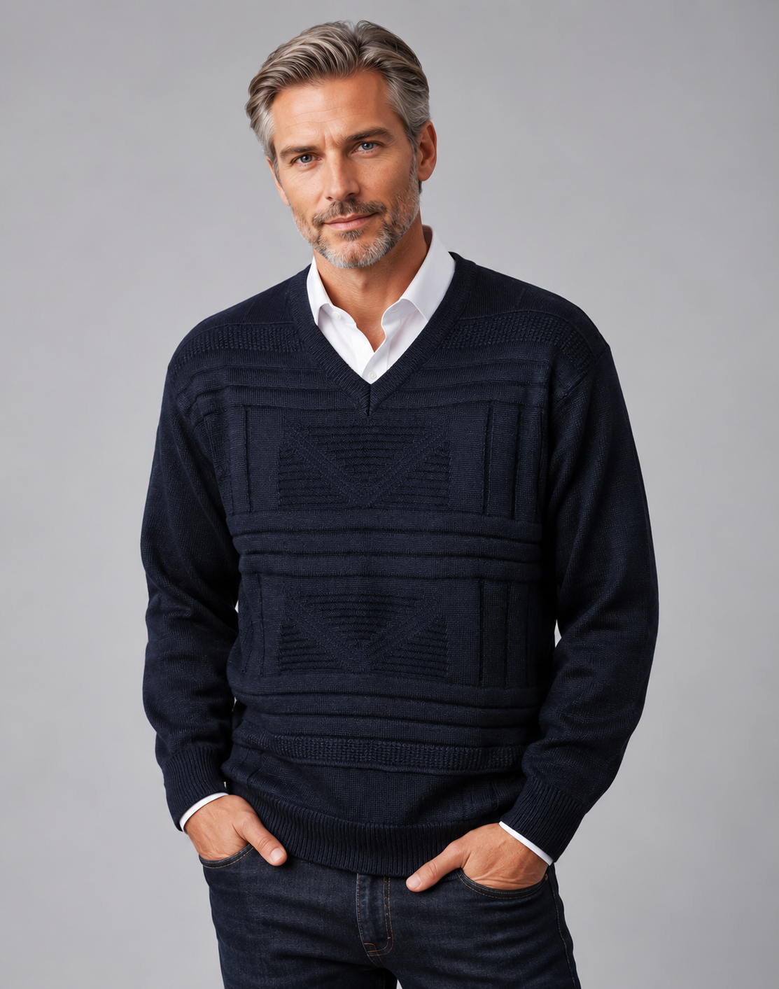 A man wearing a dark navy blue v-neck knit sweater with a geometric textured pattern, layered over a white collared shirt and dark jeans, standing against a solid gray studio background.