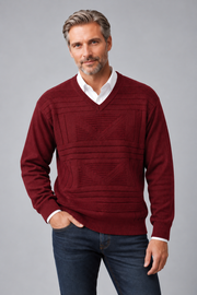 A man wearing a burgundy knit V-neck sweater with textured geometric patterns, layered over a white collared shirt and paired with dark blue jeans, against a solid gray studio background.