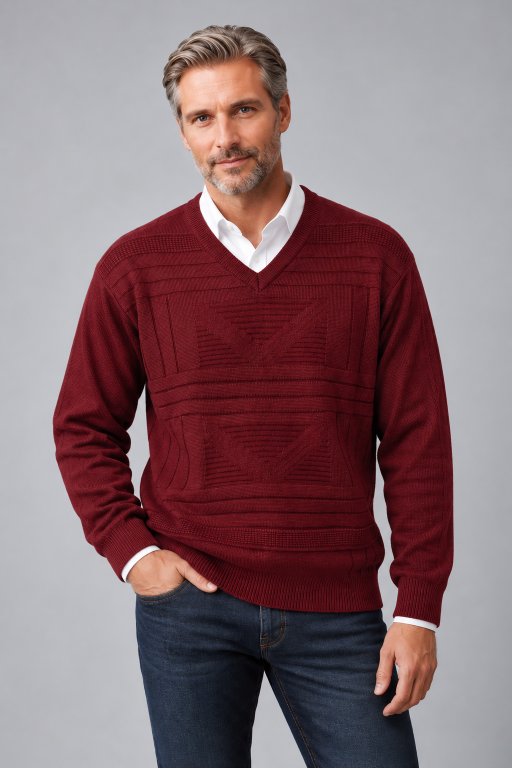 A man wearing a burgundy knit V-neck sweater with textured geometric patterns, layered over a white collared shirt and paired with dark blue jeans, against a solid gray studio background.