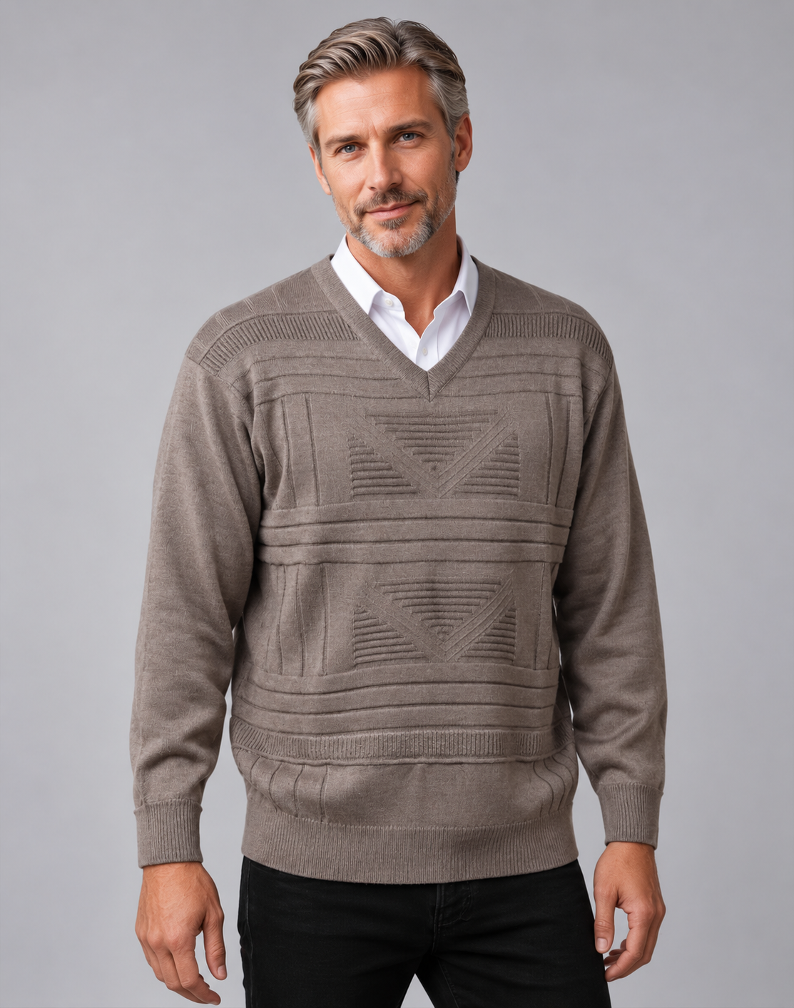 A man wearing a taupe brown knit v-neck sweater with geometric patterns over a white collared shirt and black pants, photographed against a solid gray studio background.