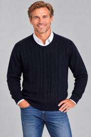 A smiling man wearing a dark navy blue cable-knit sweater over a white collared shirt and blue jeans, standing against a solid gray studio background.