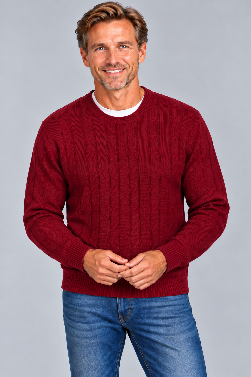 A smiling man wearing a burgundy cable-knit crewneck sweater over a white undershirt and blue jeans, standing against a solid gray studio background.