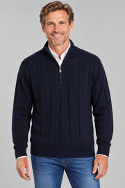 A smiling man wearing a navy blue cable-knit quarter-zip sweater over a white collared shirt and blue jeans, photographed against a plain light gray studio background.