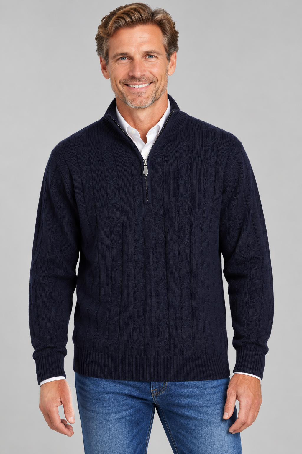 A smiling man wearing a navy blue cable-knit quarter-zip sweater over a white collared shirt and blue jeans, photographed against a plain light gray studio background.